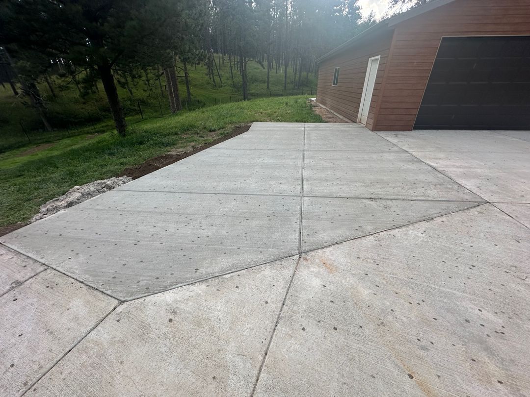 Concrete driveway beside a brick garage, bordered by grass and trees on a sloped hillside
