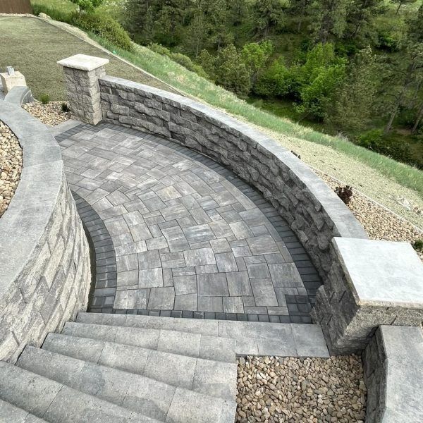 Curved stone steps leading to a paved patio area enclosed by a matching stone retaining wall, overlooking a wooded hill.