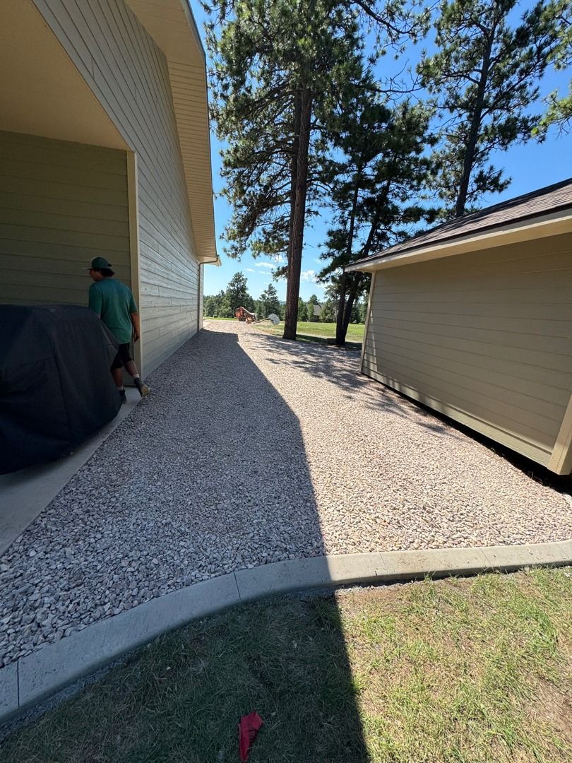 Side yard with gravel walkway, beige house, tall pine trees, and a person standing near the wall.