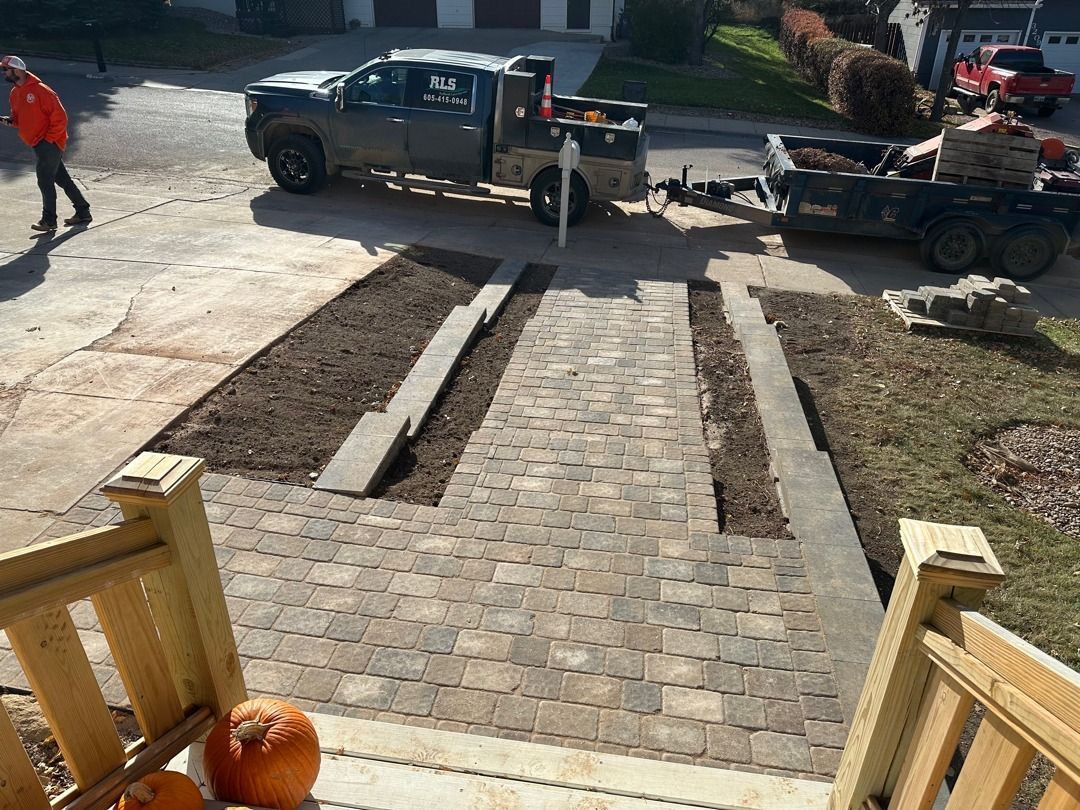 Brick walkway under construction with two pickup trucks and a worker in a driveway, viewed from a porch.