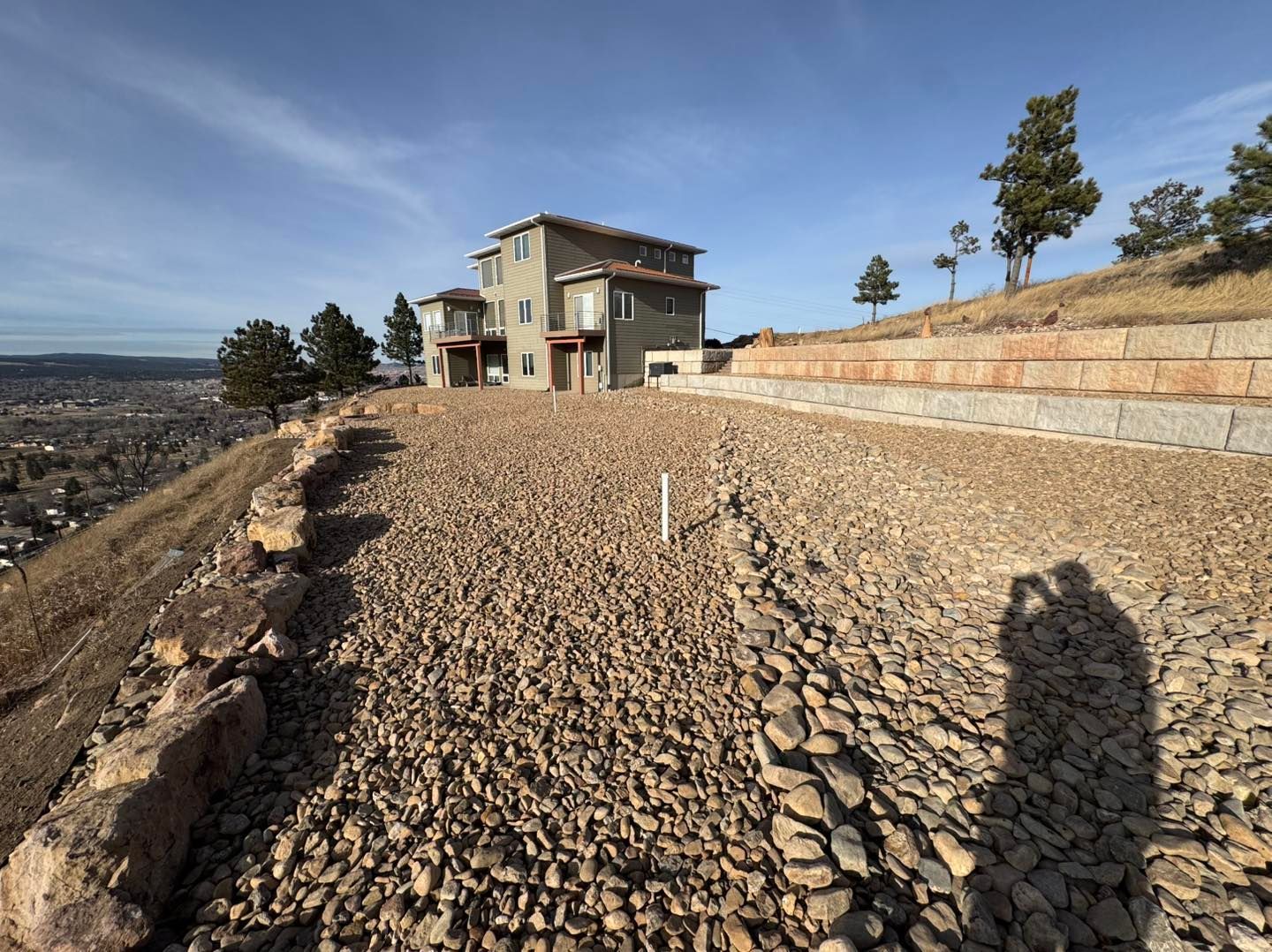 Gravel driveway leading to a house on a hillside, with stone retaining walls and a clear sky.