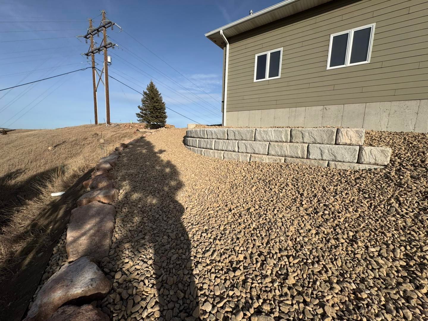 Side yard with gravel, retaining wall blocks, and a house under a clear blue sky.