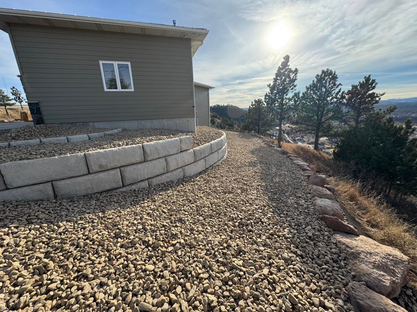Gravel path beside a gray house, with a curved stone retaining wall and trees overlooking a hillside.