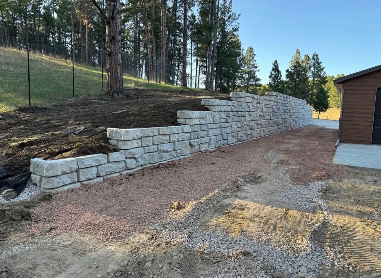 Stone retaining wall beside a gravel driveway and brown building on a wooded hillside