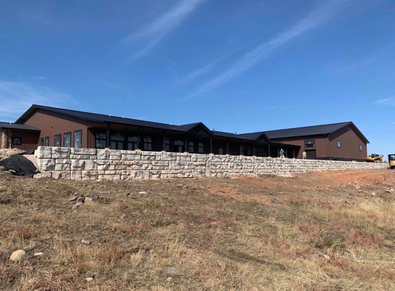 Single-story brown house with stone retaining wall on a grassy hillside under a blue sky