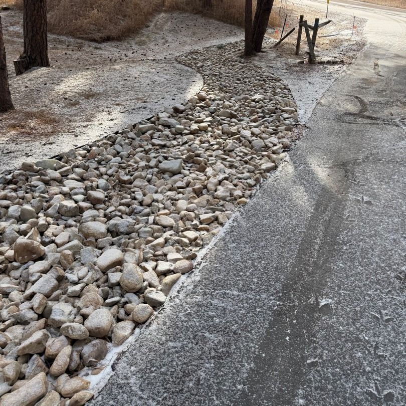 Gravel drainage swale beside a paved path in a wooded area, with rocks lining the ditch