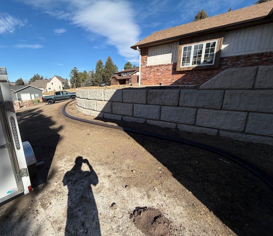 Curved retaining wall beside a house, with dirt driveway and black drainage pipe under a blue sky