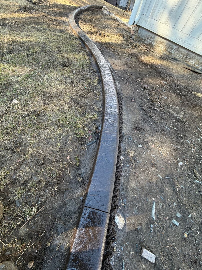 Curved black irrigation hose running along a dirt path beside a building and patchy grass.