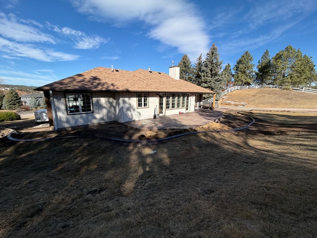 Single-story house with tan roof and white exterior in a wooded yard under a blue sky