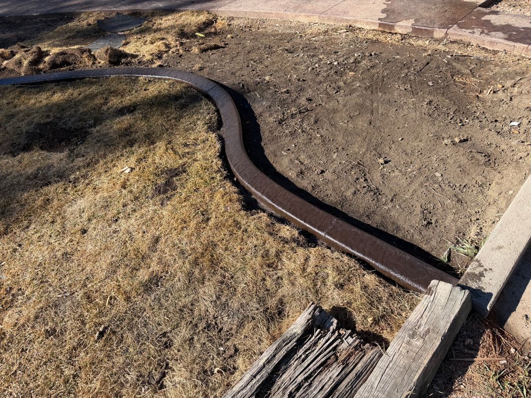 Curved rusted metal drainage channel running through dry dirt and grass beside a concrete edge