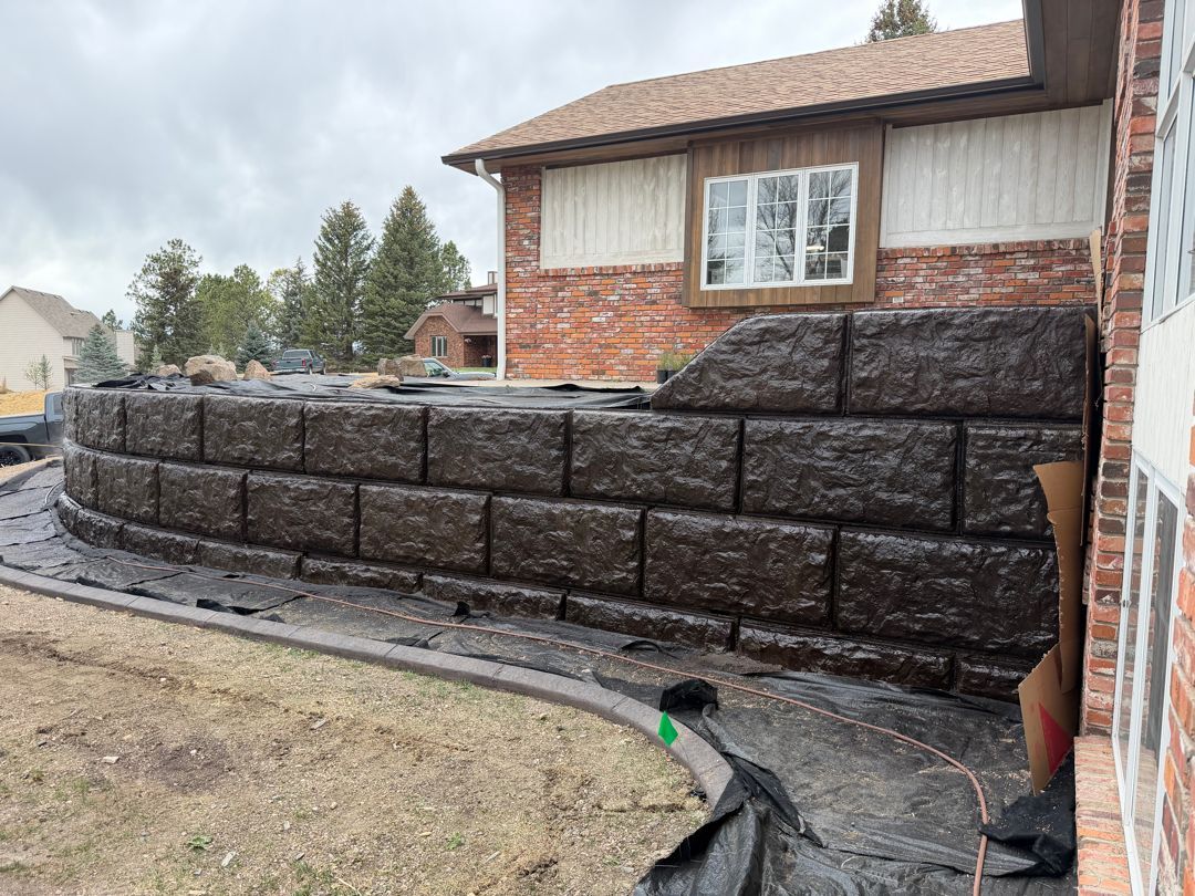 Stone retaining wall under construction beside a house, with black geotextile and gravel base.