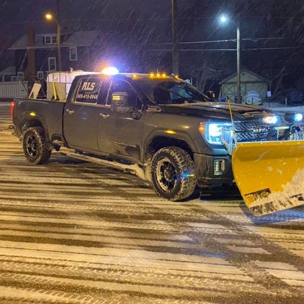 A dark pickup truck with a large yellow snowplow attached to the front driving on a snow-covered street at night.