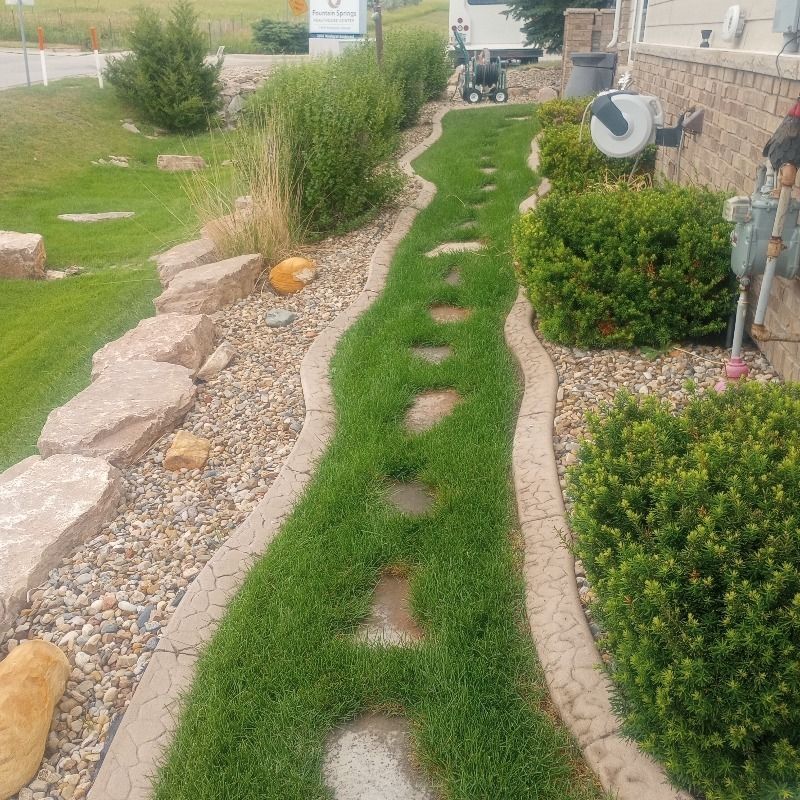 A stone path with grass-filled gaps winds through decorative gravel, lined by green shrubs and a brick building wall.