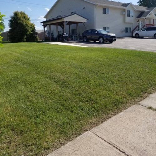 A manicured green lawn in front of a tan apartment building with a covered carport and parked cars.