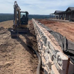An excavator sits next to a newly built stone retaining wall on a hillside construction site near a building.