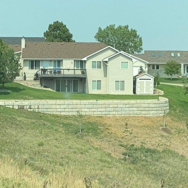 A beige suburban house with a deck and a stone retaining wall on a grassy, sloped hill under a clear blue sky.