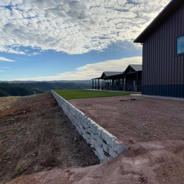 A stone retaining wall edges a gravel yard next to a modern metal-sided building overlooking a vast, hilly landscape.