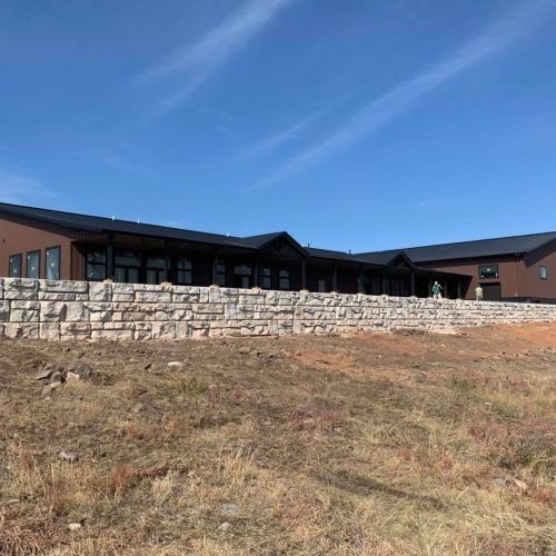 A long, dark-brown building with a black roof sits behind a tall, light-colored stone retaining wall on a grassy hillside.
