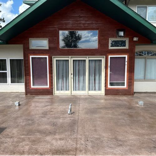 A textured patio leads to a house with a red wood front, large central glass doors, and several rectangular windows.
