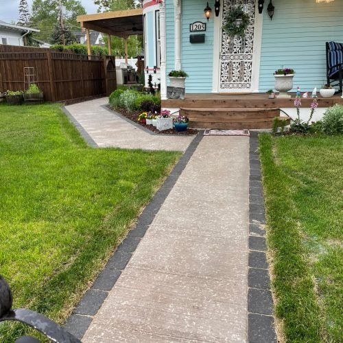 A concrete sidewalk with a dark stone border leads to the wooden front steps of a light blue house.