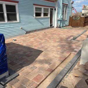 A light blue house with a newly installed multi-colored paver patio and a stone border in a backyard.
