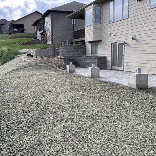 A beige house with a stone patio, retaining walls, and concrete supports, looking out over a gravel yard in a residential area.