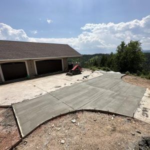 Newly poured concrete driveway extension adjacent to a house with an attached garage, set against a hilly, wooded area.