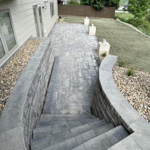 Gray stone stairs leading down to a paver patio with a curved stone retaining wall next to a house exterior.