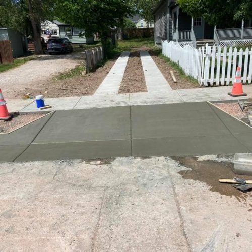 A freshly poured concrete driveway apron connects to a residential alley with two concrete tire tracks and orange cones.
