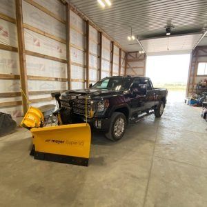 A dark-colored pickup truck with a yellow Meyer snow plow attached sits inside a bright, spacious metal-frame building.