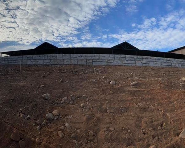 A stone retaining wall sits above a sloped, dirt embankment under a cloudy blue sky.