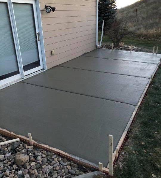 A newly poured concrete patio slab outside a house with sliding glass doors and wooden edge forms.