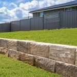A tan sandstone retaining wall lines a manicured green lawn, with a tall dark grey metal fence and house in the background.