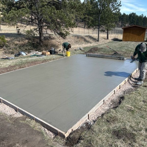 Two people finish smoothing a newly poured concrete slab surrounded by wooden forms in a grassy, outdoor setting.