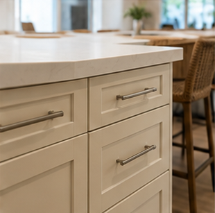 Cream kitchen island with drawers and metal handles beside a wicker barstool in a bright home kitchen