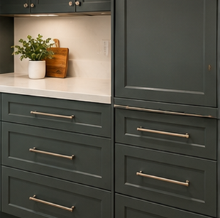 Dark green kitchen cabinets with brass handles and a white countertop with a plant and cutting board.