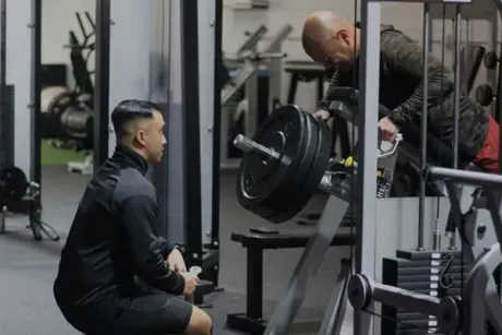 A man is squatting down in a gym while another man looks at a barbell.