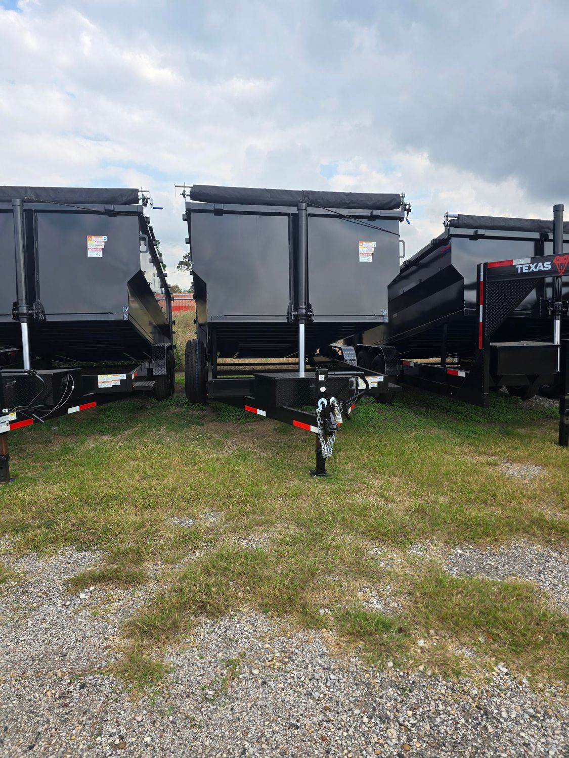 Three black dump trailers parked on grass and gravel under a cloudy sky.