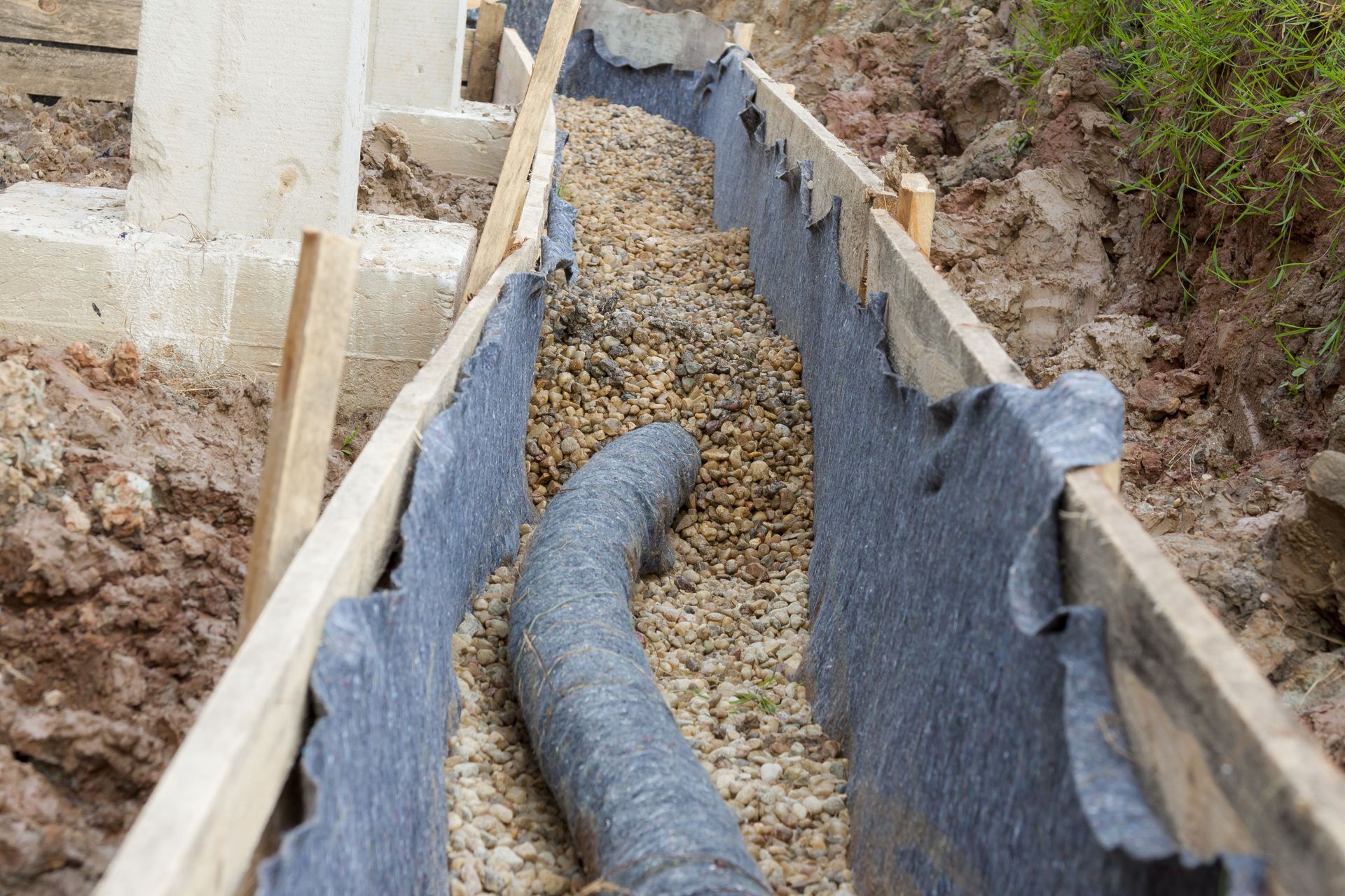 A French drain installation in a wooden-framed trench filled with gravel, lined with black landscape fabric.