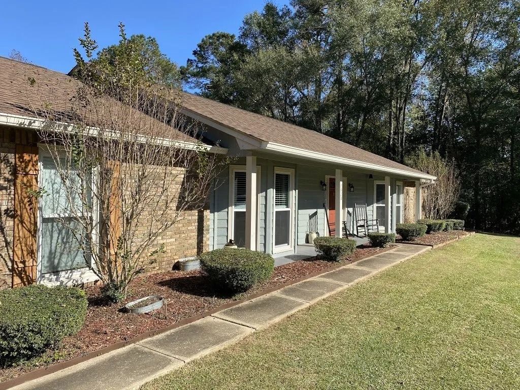 A one-story house with brick siding and a long covered porch, framed by a concrete walkway, bushes, and green trees.