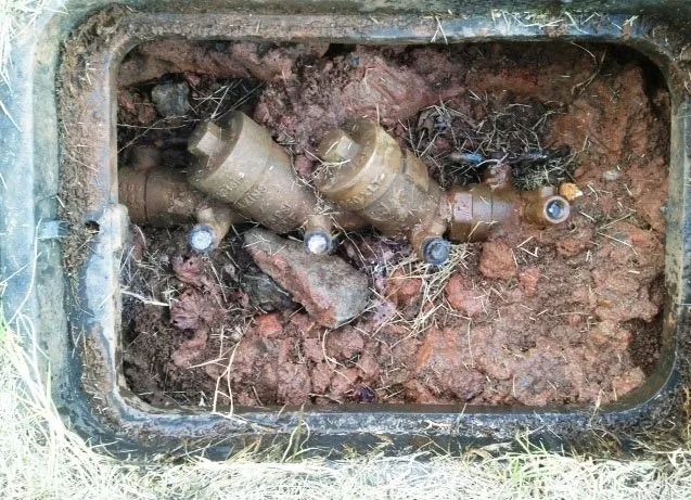 A rectangular irrigation valve box filled with dirt and debris, containing two brass sprinkler valves.