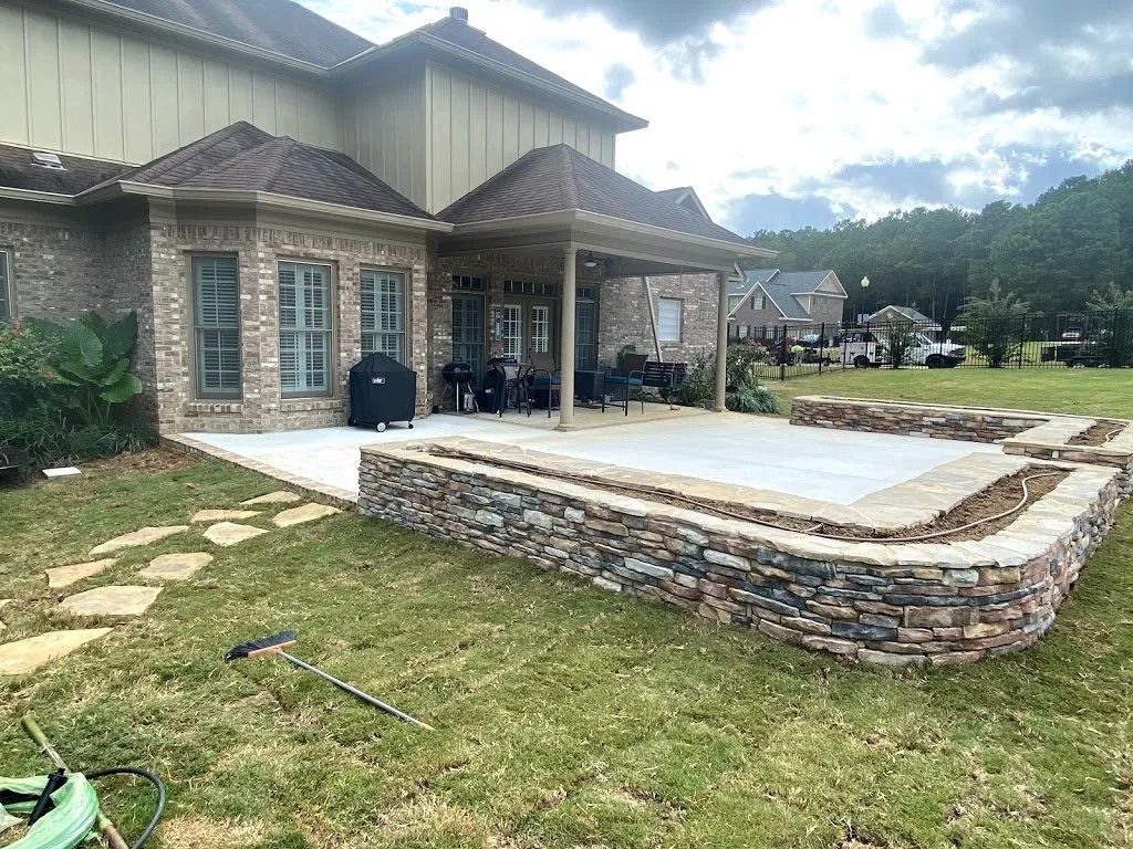 A backyard stone patio with a built-in stone retaining wall, adjacent to a brick house with a covered porch.