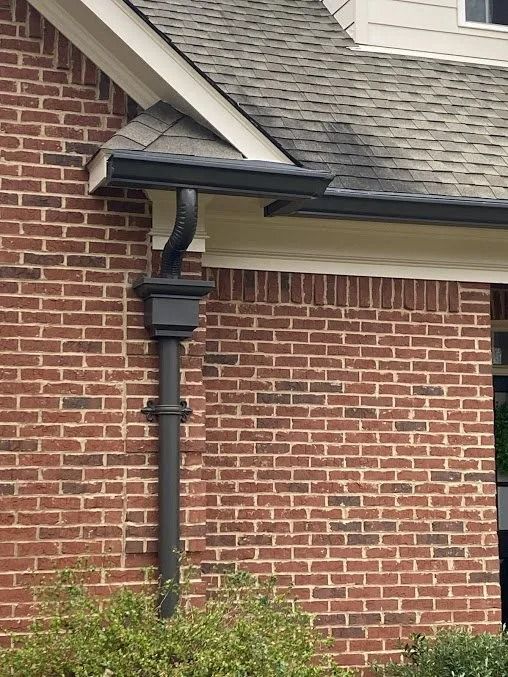A black gutter and downspout system attached to a red brick house wall under a shingled roof overhang.