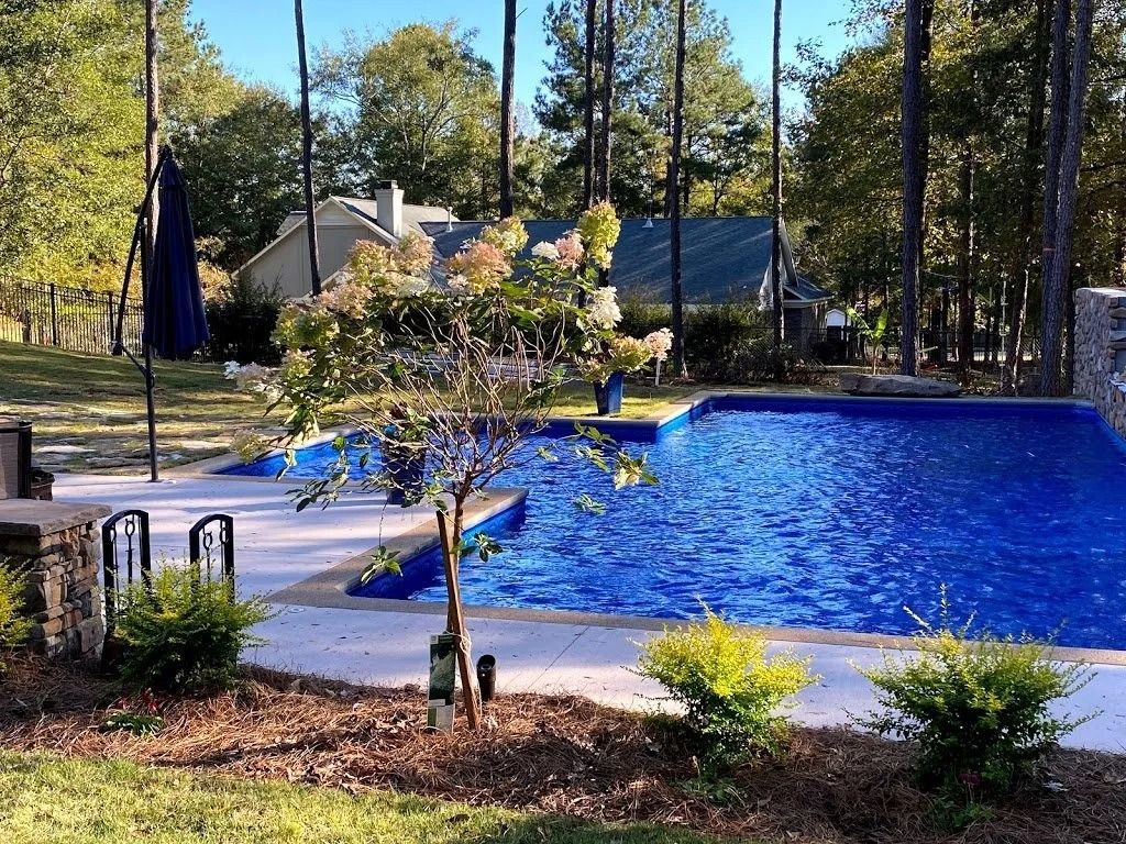 A backyard swimming pool with vibrant blue water, surrounded by trees, a patio, and small landscaping shrubs.