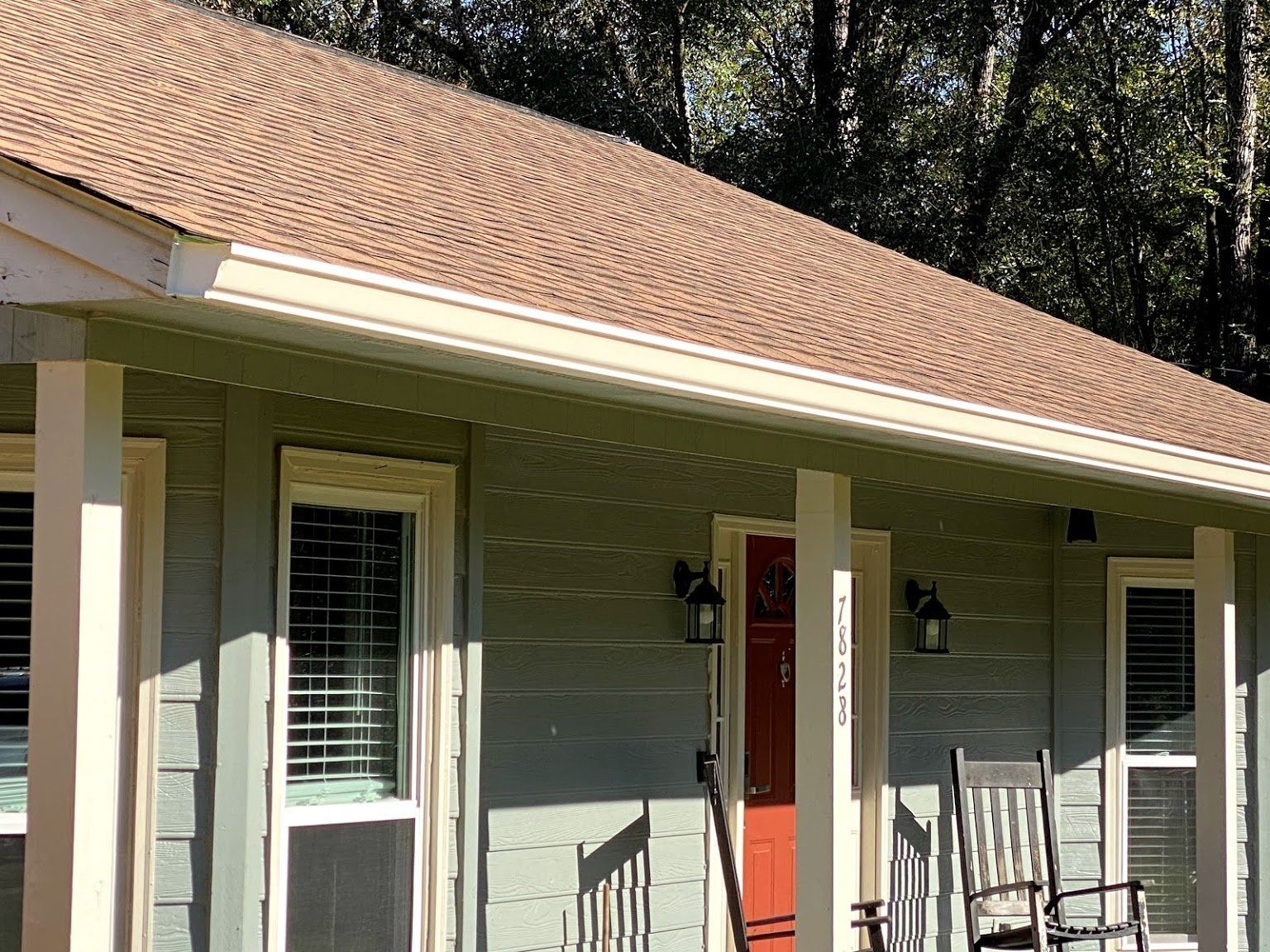 A porch with light green siding, a bright red front door, white columns, and rocking chairs under a brown shingled roof.