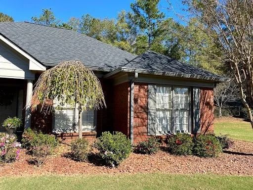 A single-story brick house with a gray shingled roof, a weeping tree in the front yard, and a green lawn under a blue sky.