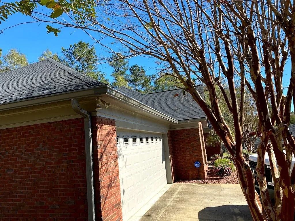 A brick house with a multi-car garage, gray shingled roof, and a bare tree in the foreground under a clear blue sky.