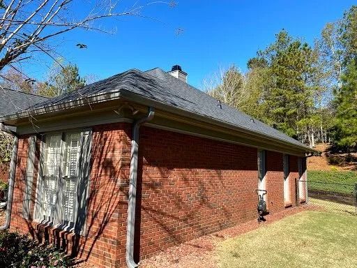 A side view of a red brick house with a dark gray shingled roof, a chimney, and a silver downspout on a sunny day.