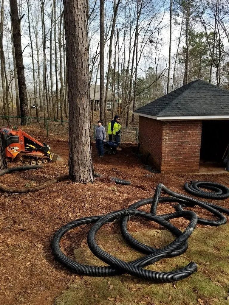Two workers in high-visibility vests stand near a brick structure and an orange skid-steer with black tubing in the yard.