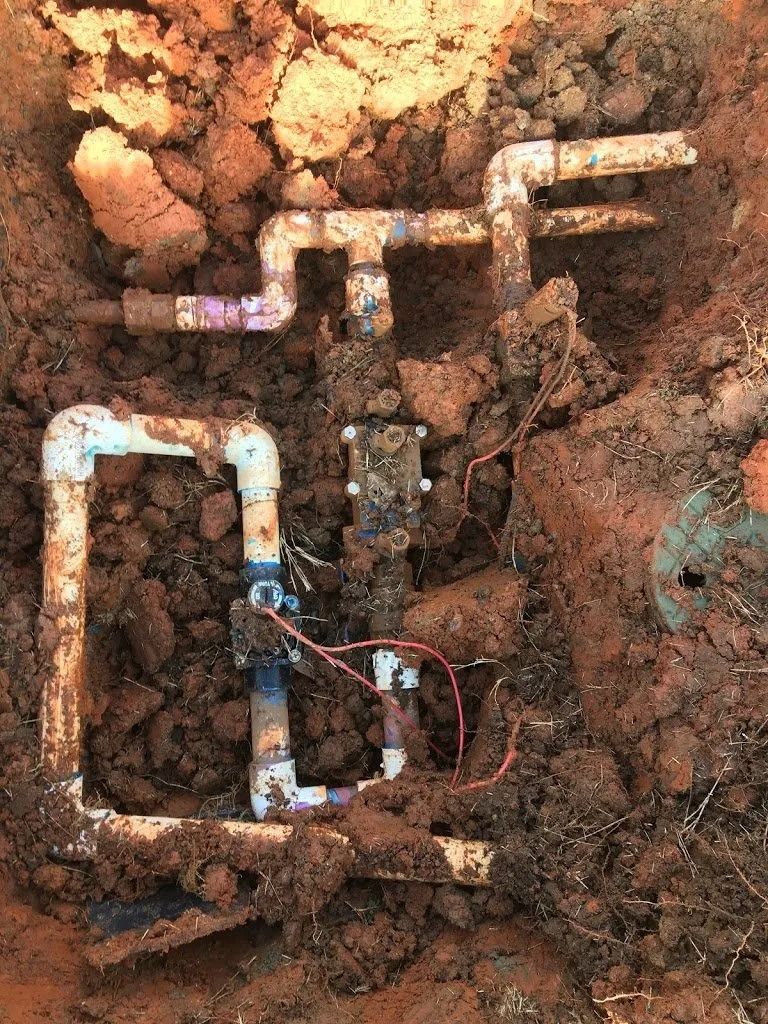 A top-down view of two irrigation valves and white PVC pipes buried in reddish-brown soil.